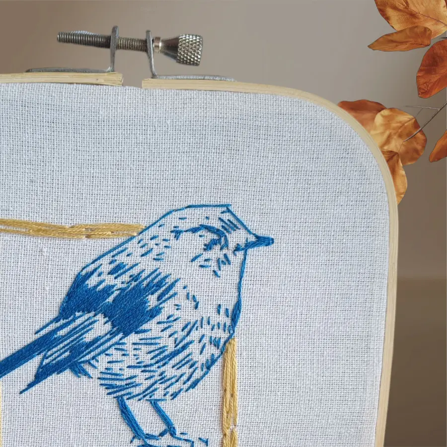 Broderie représentant un petit oiseau bleu stylisé avec une vue zoomer, posé de profil à l'intérieur d'un cadre carré doré, sur toile claire tendue dans un tambour à broder.