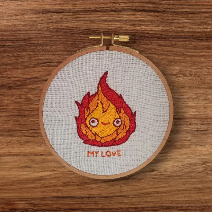 Décoration brodée à la main représentant Calcifer, personnage du Château ambulant de Ghibli, dans un tambour à broder en bois.