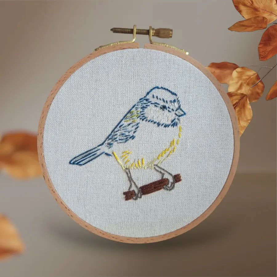 Broderie artisanale représentant une mésange bleue et jaune perchée sur une petite branche, réalisée sur toile claire et encadrée dans un tambour en bois.